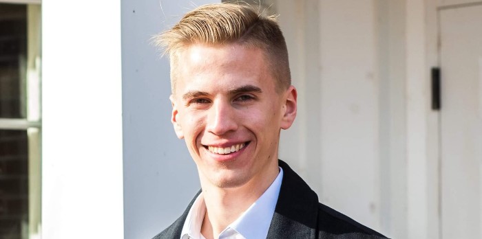 A young man with blonde hair smiles in front of a door.