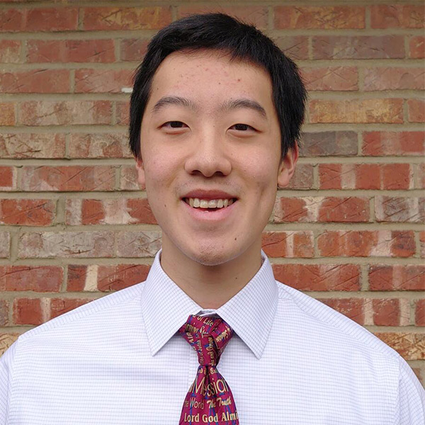 Kyle Chu stands in front of a brick wall and smiles, wearing a white shirt and red tie.