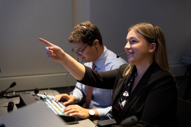 A male and female student sit at a table, wearing headsets. The woman points upwards and smiles.