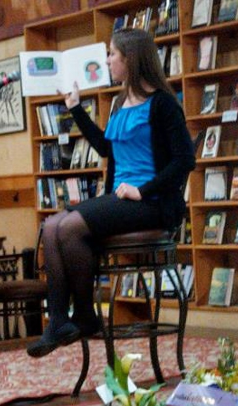 A female student sits on a tall stool holding a book open with one hand. A wooden bookshelf fills the space behind her.