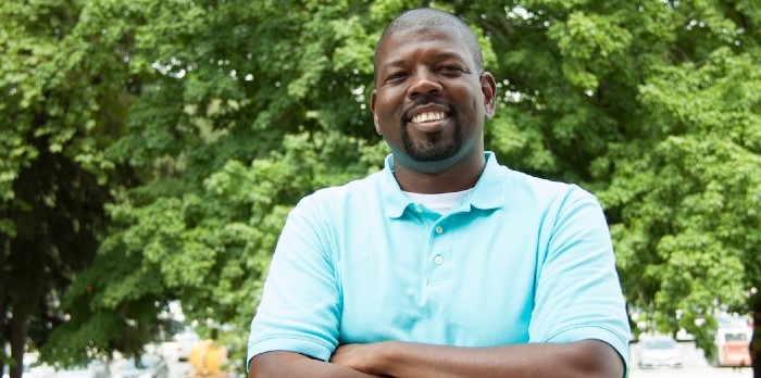 A man smiles in a collared shirt in front of a tree.