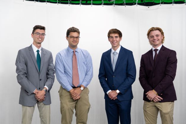 Four male students stand in front of a white backdrop, their hands clasped in front of them. They each wear a suit.