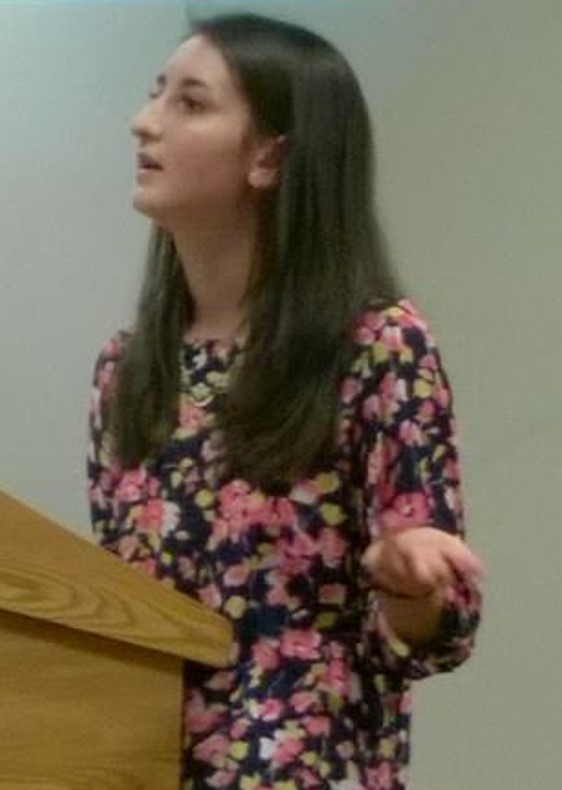 A female student with long brown hair stands behind a podium speaking
