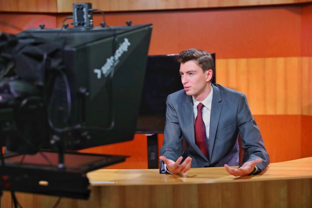 A male student in a suit sits behind an anchor desk, a camera filming him closely.