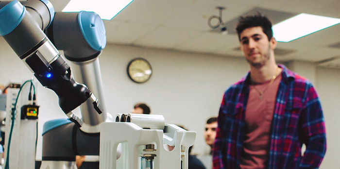 A male student stands behind a robotic arm in class.