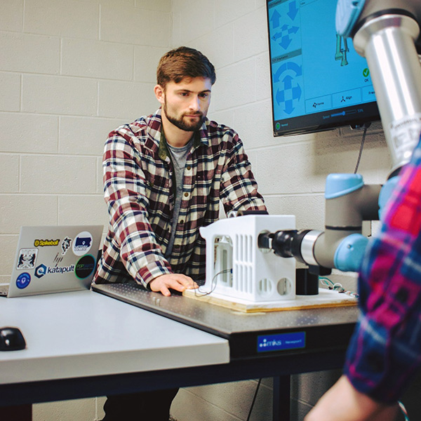 A student stands behind a table by a robotic arm and works on a laptop.