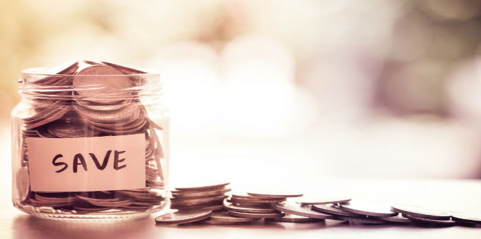 There is jar filled with coins inside and some spilling over the side. There is a white piece of paper on the jar that says "save" on it in black letters.