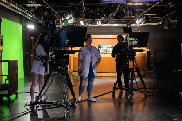 Three students stand in a studio behind two cameras, a green screen to their left.