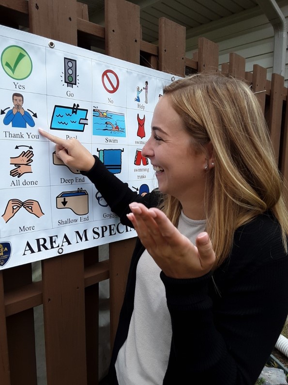 A female student stands sideways and points to a white poster beside her with small colorful graphics.