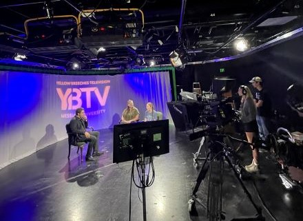 Three students sit in front of a sheet with the YBTV logo projected onto it. Two more students stand to the side and film them behind a camera.