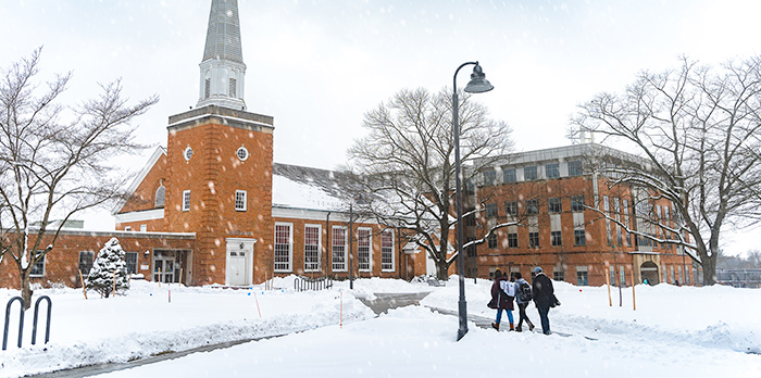 Three students walk down the sidewalk towards Hostetter Chapel. Snow covers the grass and falls from the sky.