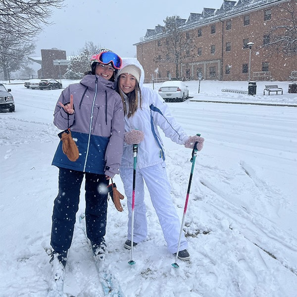 two female students in winter gear stand outside in the snow wearing skis.