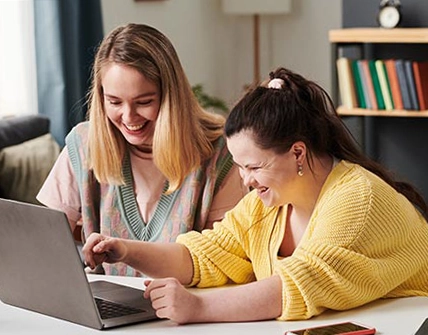 A woman and a student sit in front of a laptop, reading something on the screen. Both are smiling. 