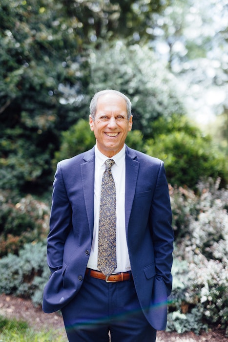 Jon C. Stuckey in a navy suit and patterned tie standing outdoors in a garden setting.