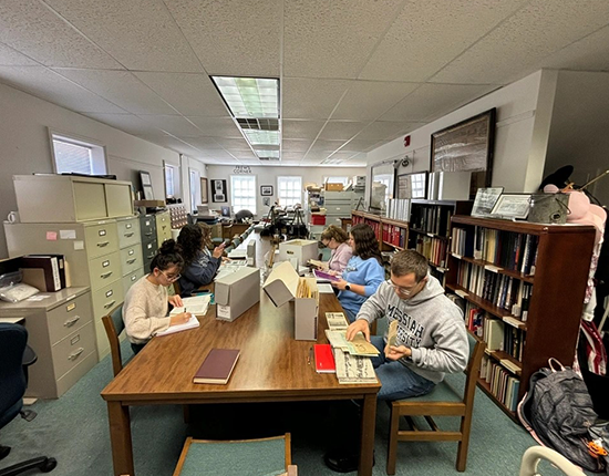 Students sit around large wooden table and look through folders.