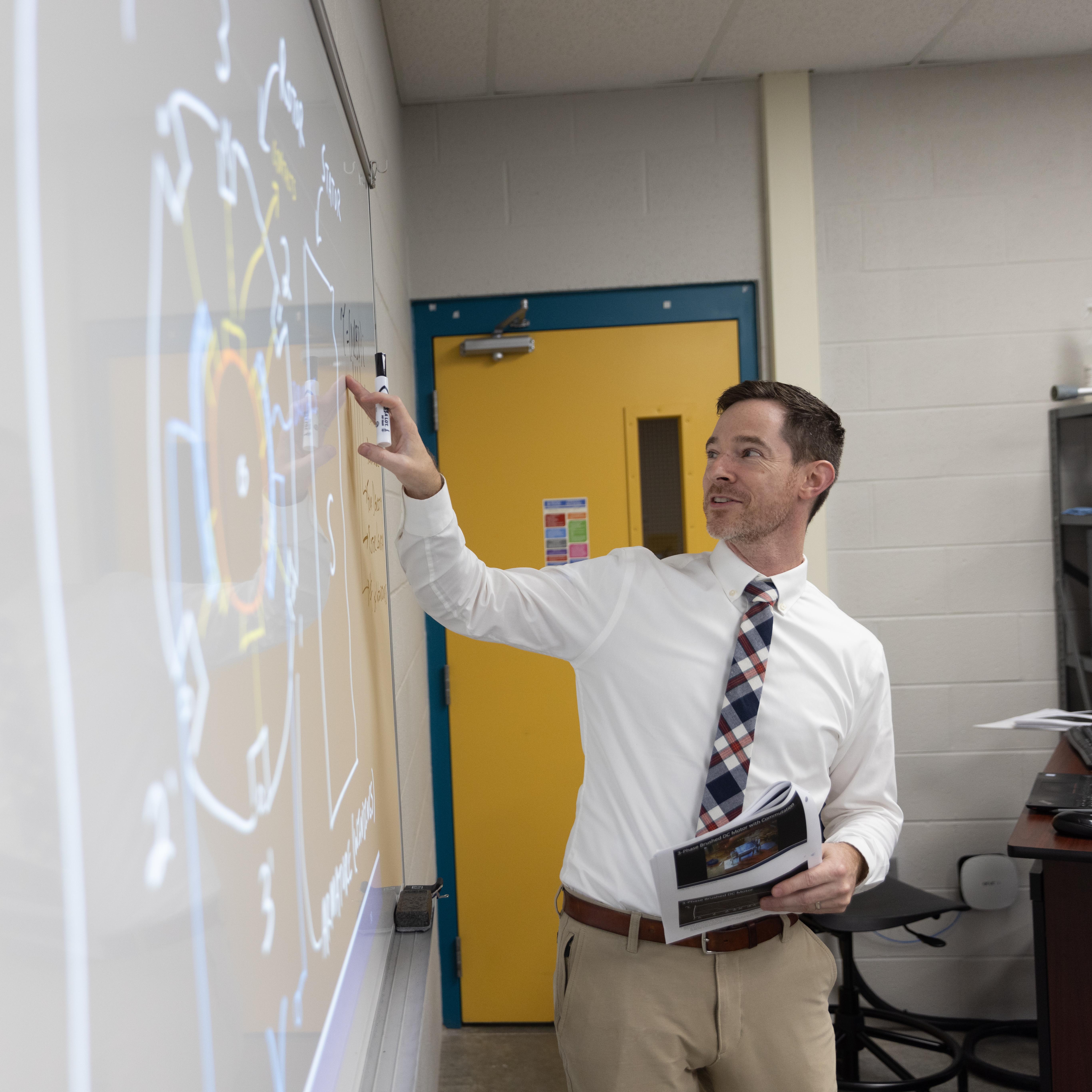 A male teacher stands at a whiteboard, instructing a class.