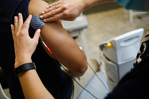 A woman holds a device against another person's bicep, cords extending out of frame.
