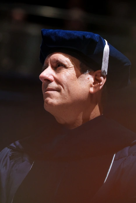 Close-up profile of President Stuckey wearing academic regalia and a blue doctoral tam with a white tassel, lit from the side against a dark background.