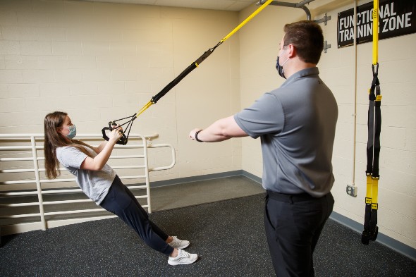 A female student in an athletic training facility does pull-ups on ropes while an instructor watches.
