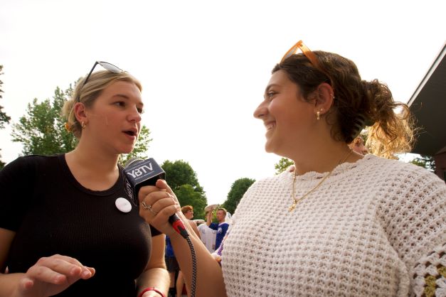 Two female students stand outside. The one of the right holds a microphone to the one on the left, who speaks into it.