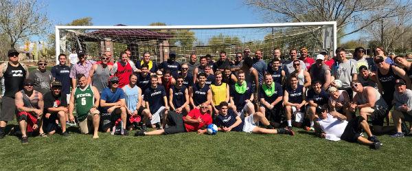 A large group of soccer players, some old and some young, stand in front of a goal smiling for the camera.