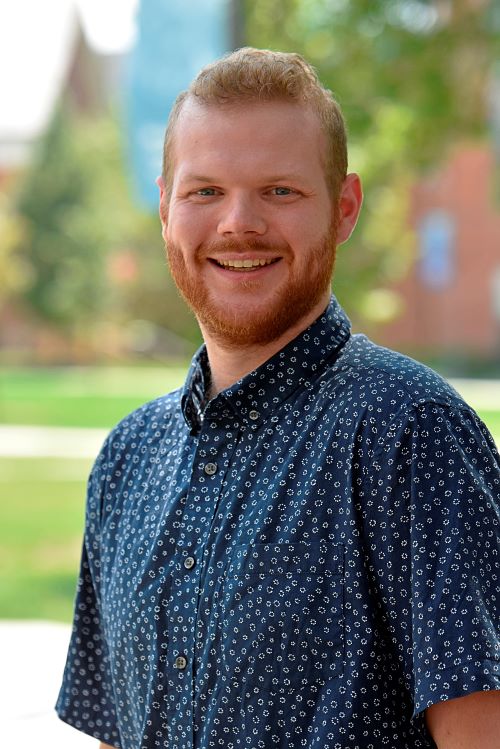 Smiling young man with a beard stands outdoors in a casual shirt, posing in front of a brick building with windows.