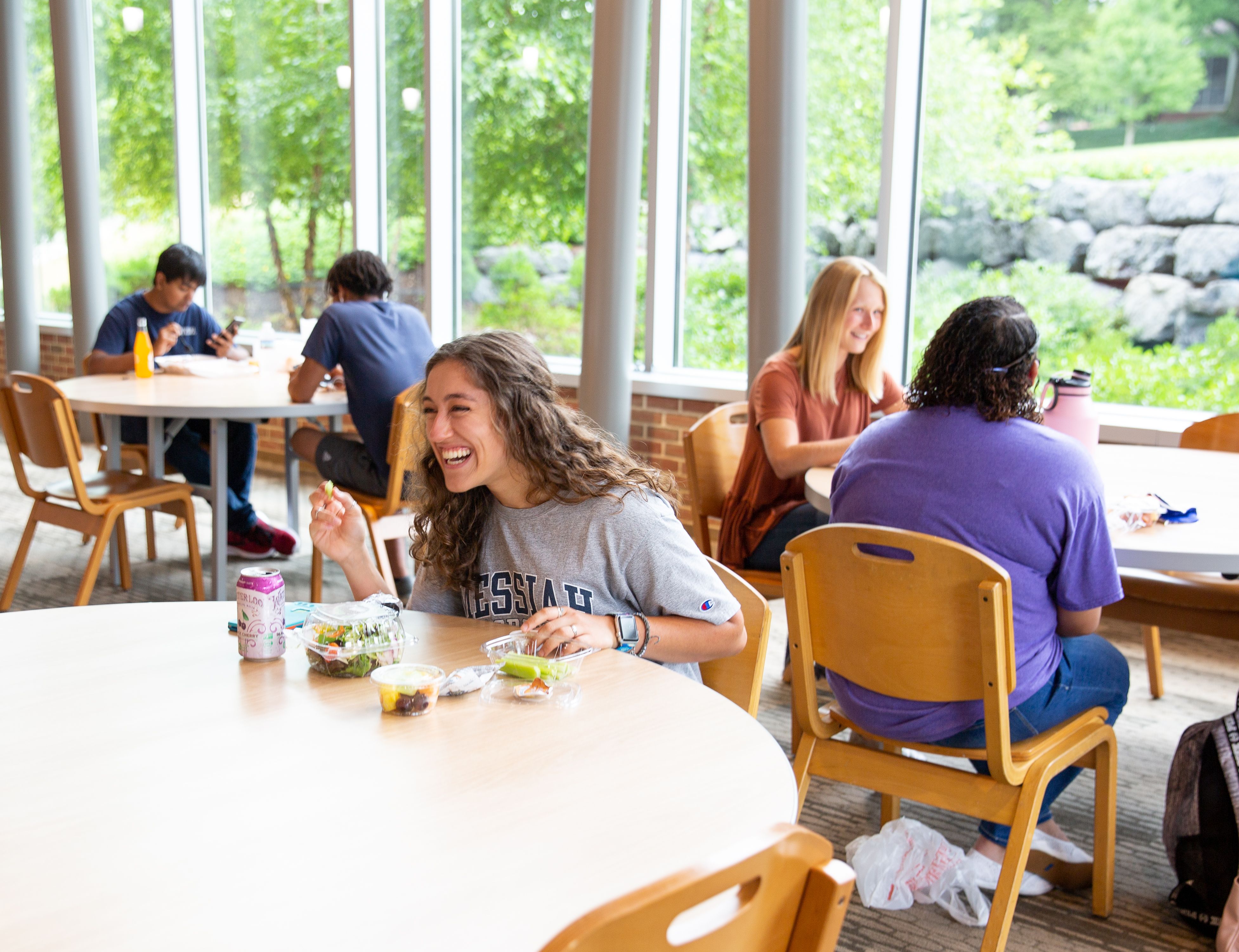 A student enjoys lunch with a salad at a bright, spacious dining area filled with other students socializing.