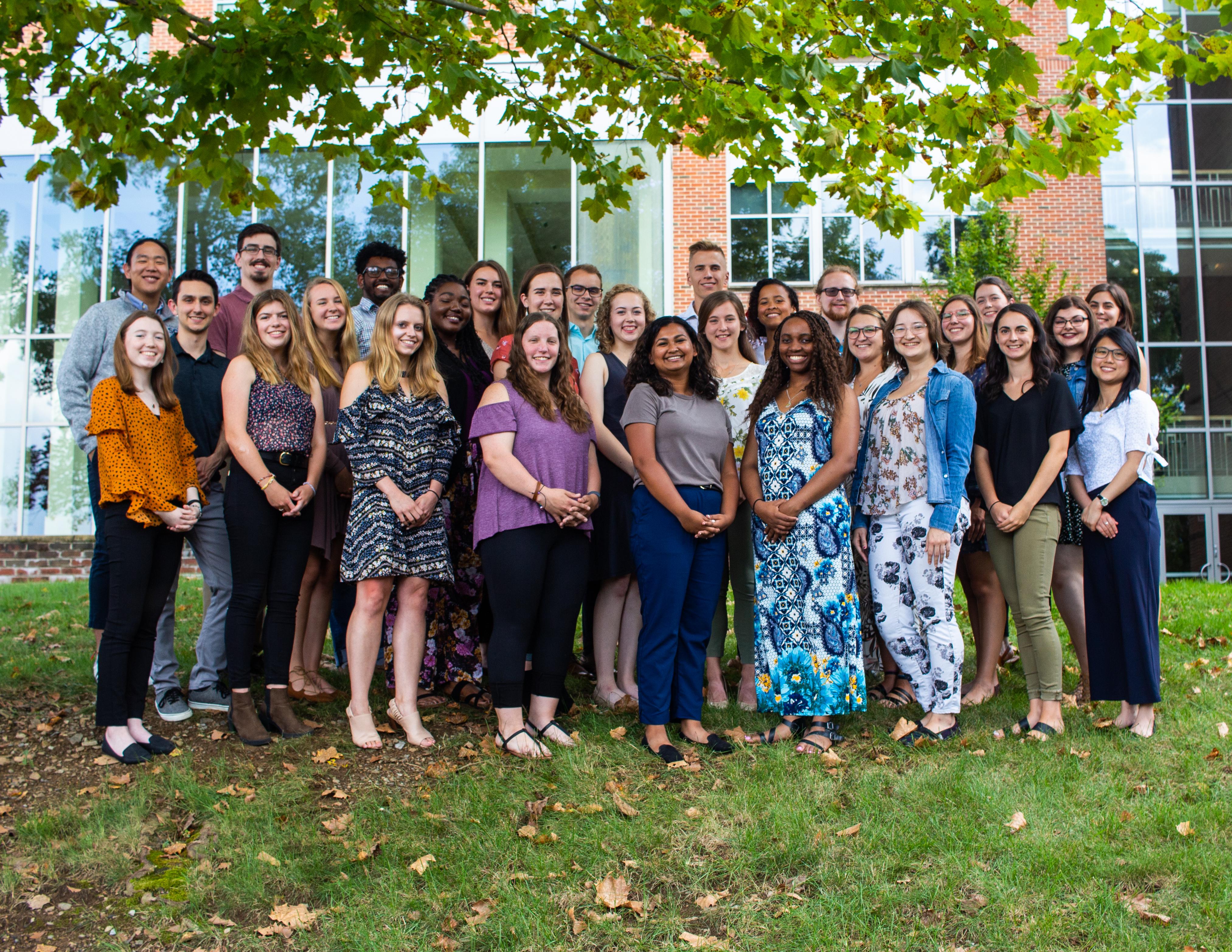 A diverse group of young professionals poses together outdoors in front of a modern building, surrounded by green grass and trees.