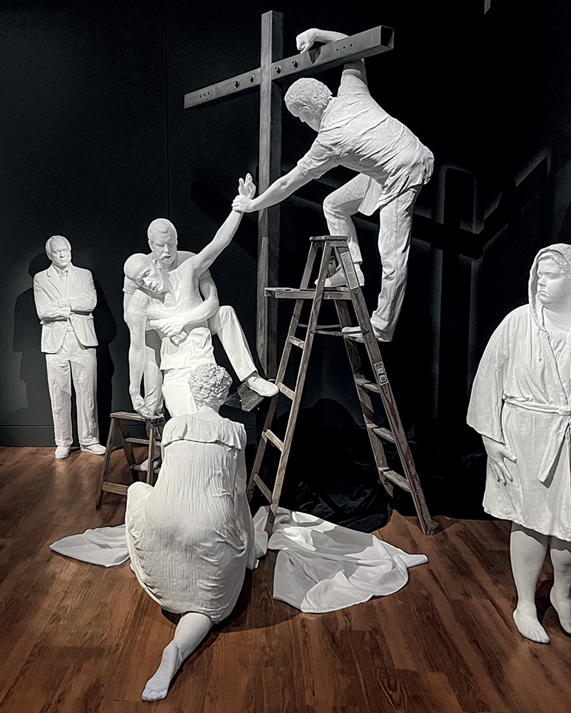 A white sculpture installation depicting figures in a dramatic scene, including a man on a ladder reaching towards another figure, while a kneeling woman and other figures observe.