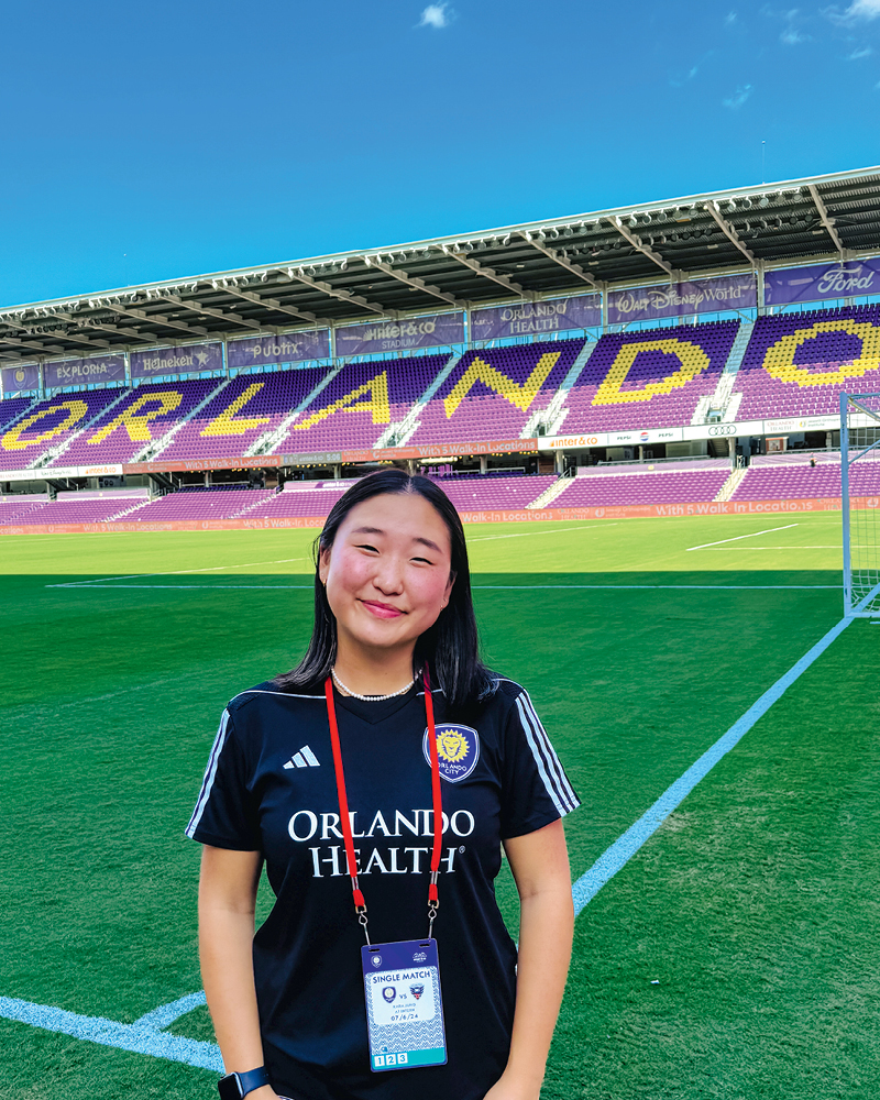 Young woman wearing an Orlando City SC jersey stands on the soccer field, with the stadium seating displaying "ORLANDO" in the background.