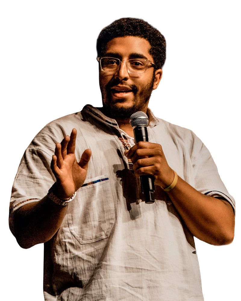 A young man with glasses speaks into a microphone while wearing a light-colored, traditional-style shirt.