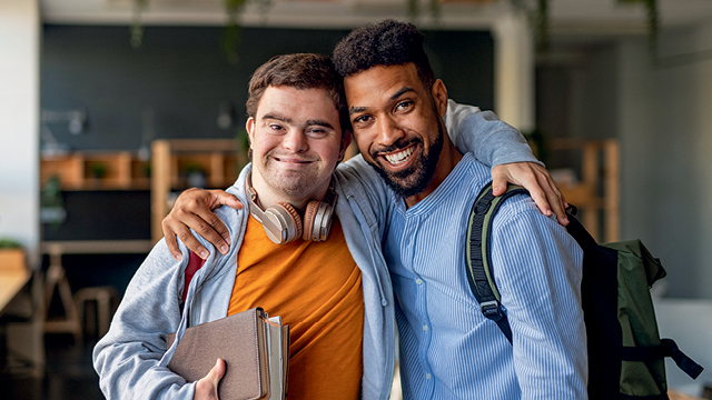 Two smiling young men, one with Down syndrome and the other with a beard, embracing each other in a modern, bright study space. Both are wearing casual clothing, with one holding books and wearing headphones.