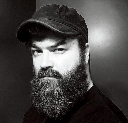 A black and white portrait of Benjamin Jancewicz with a full beard and a cap, gazing intently at the camera.