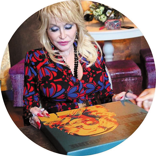 Dolly Parton signing a book with a colorful cover illustration, wearing a vibrant patterned outfit.