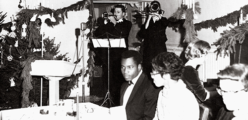 Black and white photo of a 1960s holiday party featuring a trumpet player and a singer performing, with guests seated at tables decorated for the occasion.
