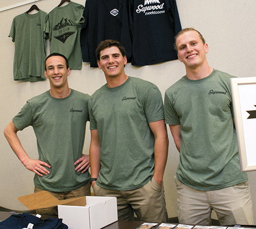 Three young men wearing matching green shirts stand behind a table showcasing merchandise for the Sapwood brand.