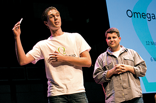 Two young men presenting on stage, one holding a pen and gesturing while the other stands beside him with a thoughtful expression.