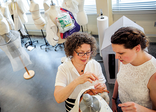 A fashion design instructor guides a student in a studio filled with dress forms and materials.