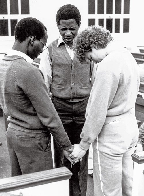 Powerhouse then and now, older photo, singers standing, holding hands in prayer in Hostetter chapel.