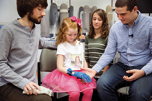A child with a blue prosthetic arm interacts with two adults, showcasing advancements in assistive technology.