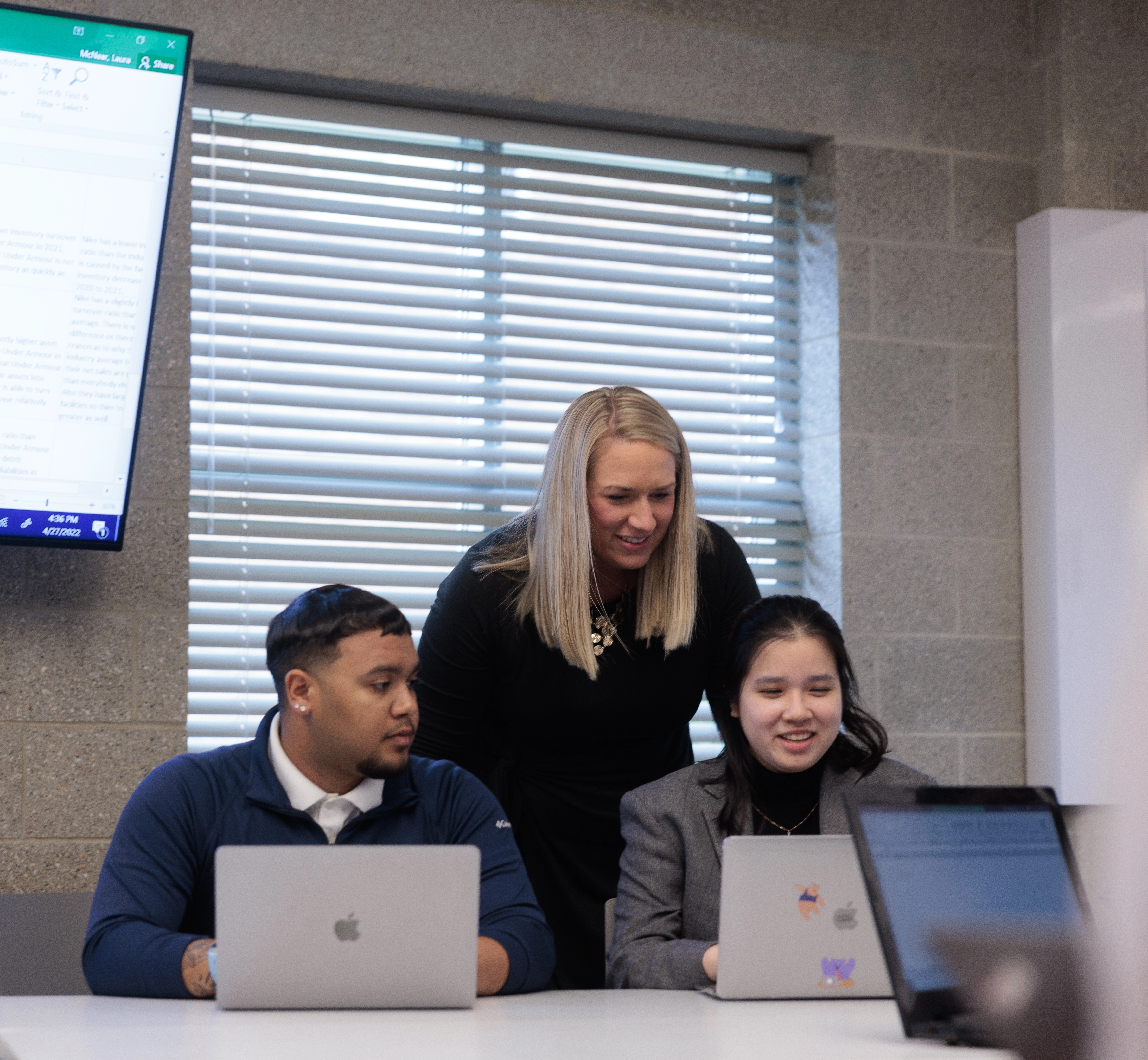 A female professor stands behind 2 students, who are sitting at open laptops.