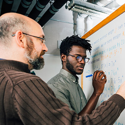 A student writes mathematical equations on a whiteboard while an instructor stands beside him, observing and offering guidance in a classroom setting.