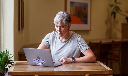 A woman working on a laptop at a wooden table in a cozy indoor setting.