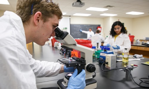 A student examines a sample under a microscope in a laboratory setting, while another student conducts an experiment in the background.