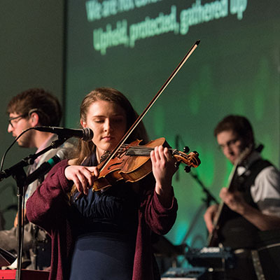 A woman plays the violin on stage alongside other musicians during a live performance, with song lyrics projected on the wall behind them.