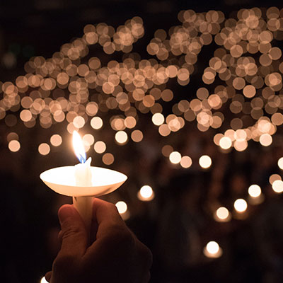 A hand holds a lit candle in a dark room filled with many other glowing candles held by people in the background.