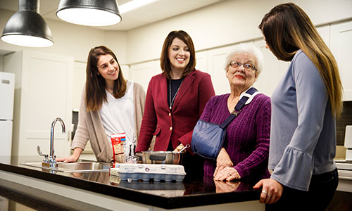 A group of four women, including an elderly woman with a shoulder sling, interact in a modern kitchen setting, emphasizing community and support.