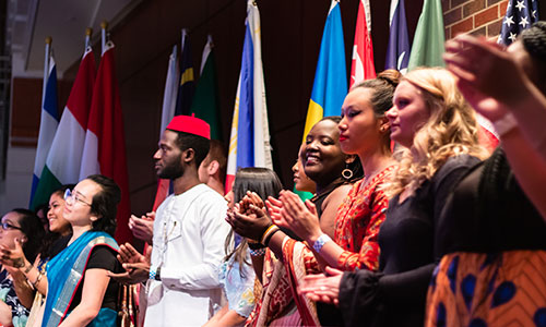 A diverse group of individuals standing together, showcasing cultural attire and clapping in front of flags from various countries.